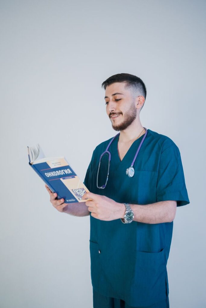 Young male nurse reading an oncology book in scrubs with a stethoscope in a bright setting.