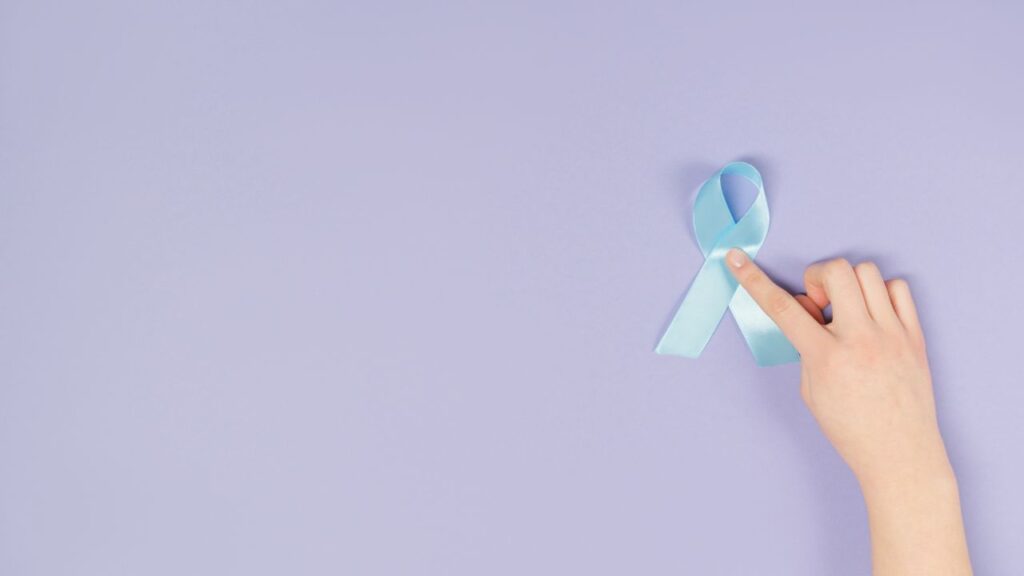 A hand holding a blue awareness ribbon on a light purple background symbolizing support and awareness.