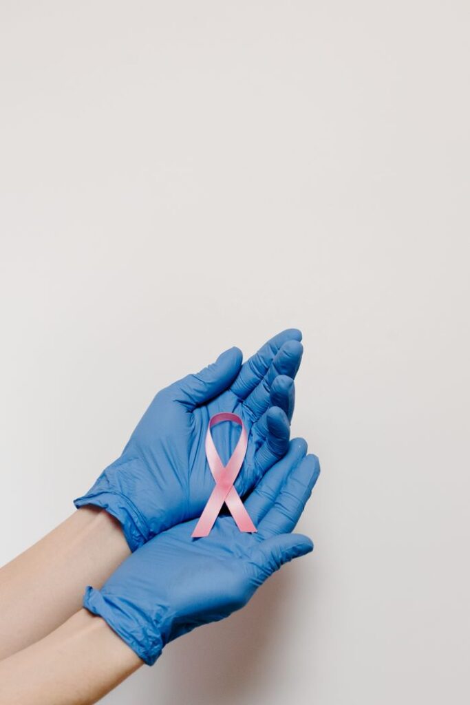 A pair of hands in blue gloves holding a pink cancer awareness ribbon against a white background.